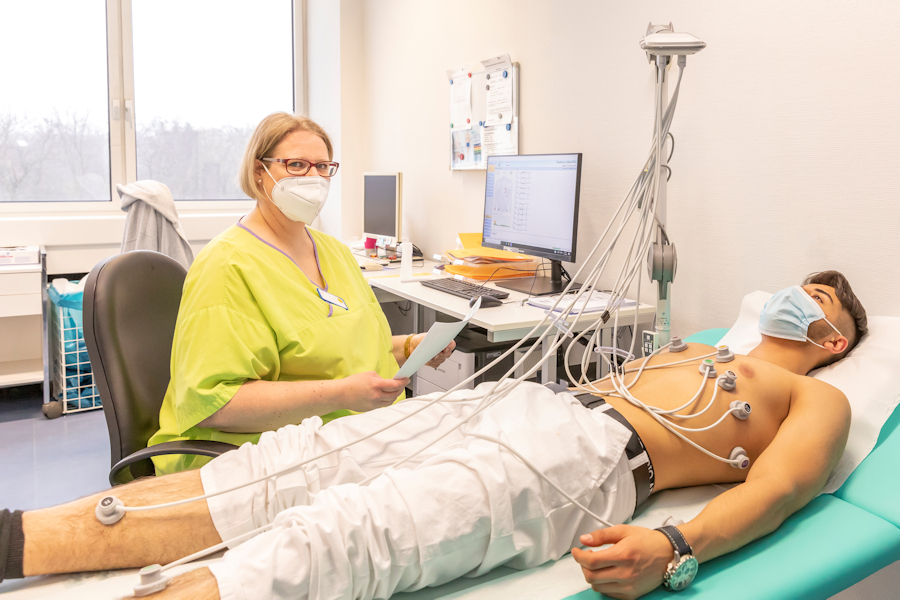 Sitzende Gesundheits- und Krankenpflegerin betreut einen liegendem Rehabilitanden an dessen Körper Dioden kleben bei der EKG Aufzeichnung 